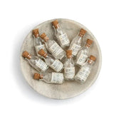 A top-down view of a round, light gray ceramic plate or tray holding several small clear glass vials with cork stoppers and white labels, set against a plain white background.